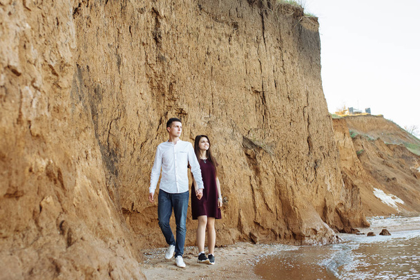 young, happy, loving couple, near the sand wall, on the sea, holding hands go and look at each other, - Фото, зображення