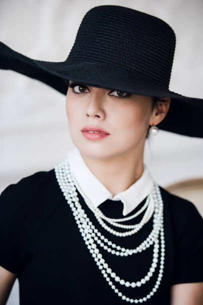 Attractive woman wearing black dress, hat and pearls, sitting on chair - Photo, image