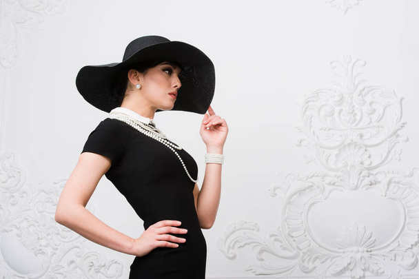 Portrait of a beautiful young woman in retro style in an elegant black hat and dress over luxury rococco wall background - Fotoğraf, Görsel