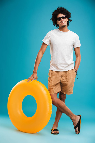 Photo of happy handsome young african curly man standing isolated over blue background looking camera holding rubber ring. - Фото, зображення