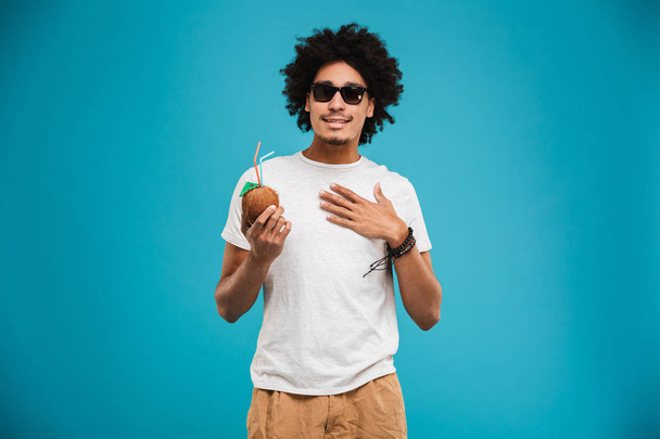 Photo of handsome happy young african curly man standing isolated over blue background drinking cocktail coconut. - Foto, imagen