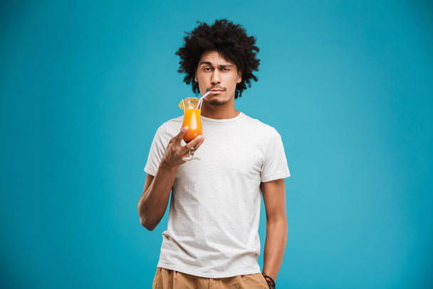 Portrait of a confident young african man drinking tropical cocktail isolated over blue background - Foto, immagini