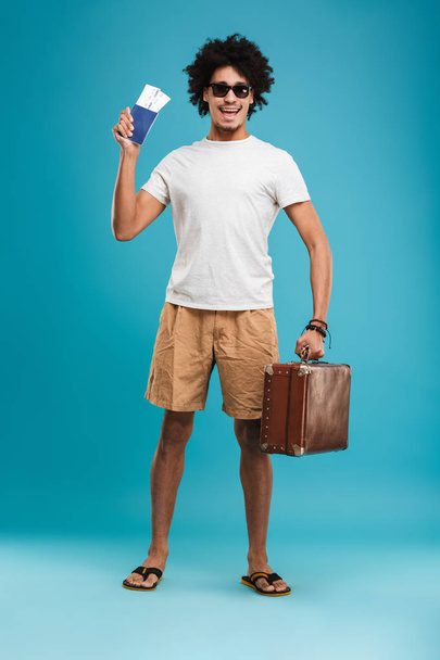 Picture of handsome happy young african curly man tourist isolated over blue wall background holding tickets with passport and suitcase. - Fotó, kép