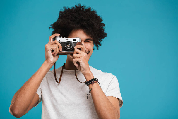 Picture of handsome emotional young african curly man photographer isolated over blue wall background holding camera. - Φωτογραφία, εικόνα