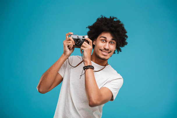 Picture of handsome emotional young african curly man photographer isolated over blue wall background holding camera. - Foto, afbeelding