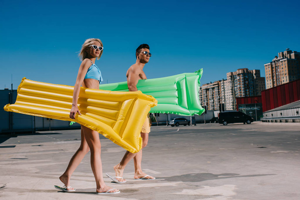 attractive young couple in beach clothes with inflatable beds walking on parking - Фото, изображение