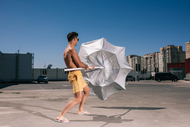 красивый молодой мужчина без рубашки с пляжным зонтиком на парковке
 - Фото, изображение