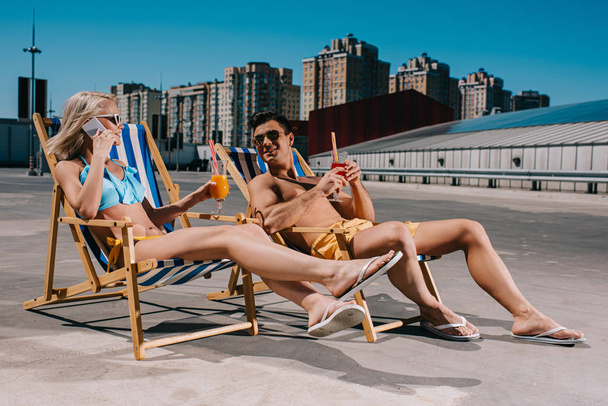 young couple with glasses of cocktails while sitting on sun loungers on parking - Photo, Image