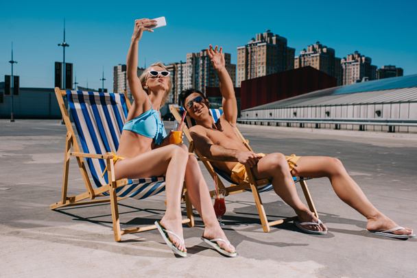 young couple relaxing on sun loungers with cocktails and taking selfie on parking - Photo, Image