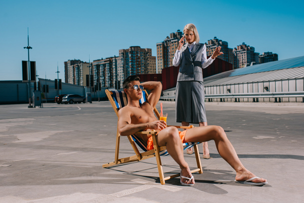 красивий молодий чоловік розслабляється на сонці в той час як його божевільна леді бос в костюмі стоїть поруч з ним і розмовляє по телефону на парковці
 - Фото, зображення