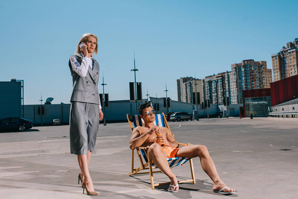 attractive young man relaxing on sun lounger while his lady boss in suit next to him and talking by phone on parking - Zdjęcie, obraz