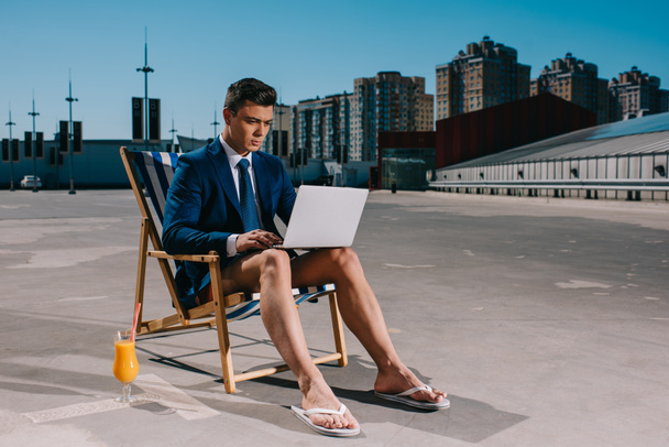 joven hombre de negocios guapo en pantalones cortos y chaqueta de trabajo con el ordenador portátil mientras está sentado en la tumbona en el aparcamiento
 - Foto, Imagen