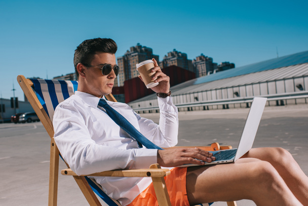 handsome young businessman in shorts with coffee to go working with laptop while sitting on sun lounger on parking - Photo, image