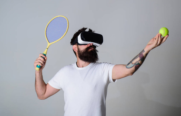 Man with beard in VR glasses, grey background. Hipster on concentrated face use modern technologies for sporty entertainment. VR tennis concept. Guy with VR glasses play tennis with racket and ball - Valokuva, kuva