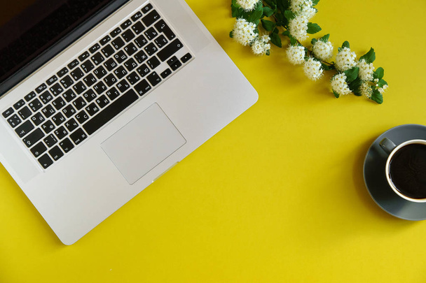laptop with hot coffee in cup and branch with flowers on bright olive yellow background, concept of female blogger workspace  - Φωτογραφία, εικόνα