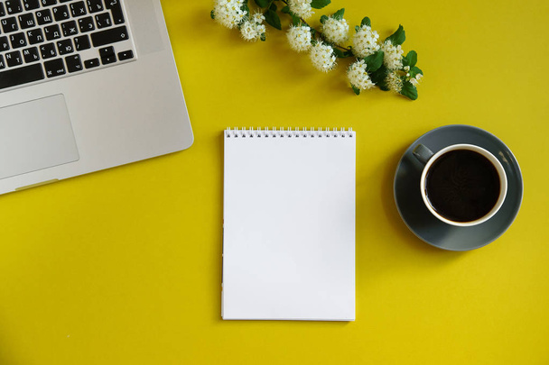 laptop with hot coffee cup and branch with flowers and notebook on bright olive yellow background, concept of female blogger workspace  - Photo, Image