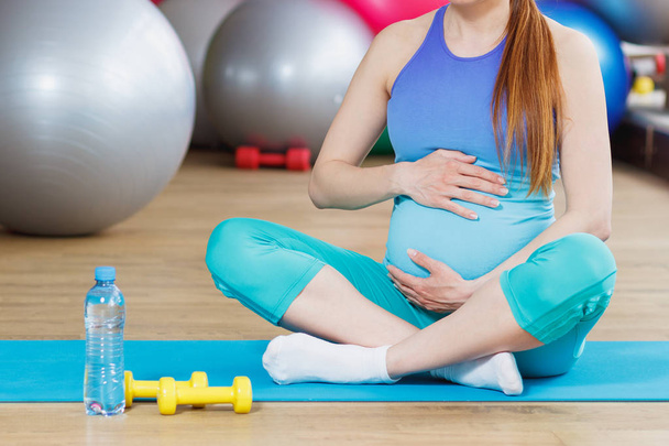 Mulher grávida senta-se no tapete no ginásio, fundo foco suave
 - Foto, Imagem