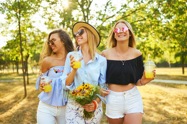 Три гарні дівчата розважаються і п'ють жовтий сік у парку. Відмінна сонячна погода. Прекрасні фігури. Супер настрій. Літня концепція
. - Фото, зображення