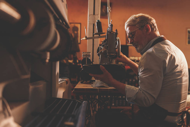 Un zapatero de edad avanzada en la máquina de la fábrica en interiores
 - Foto, imagen