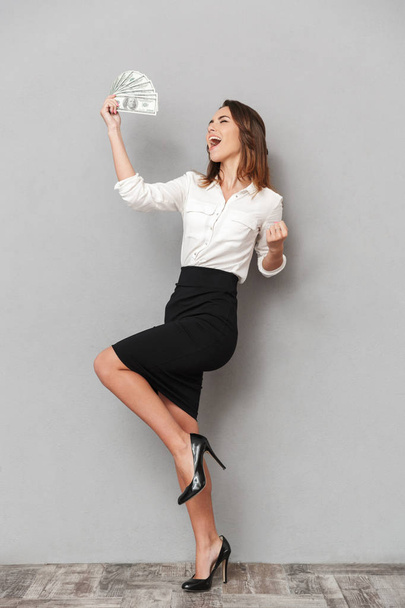 Photo of excited young business woman standing isolated over grey wall background make winner gesture holding money. - Fotó, kép