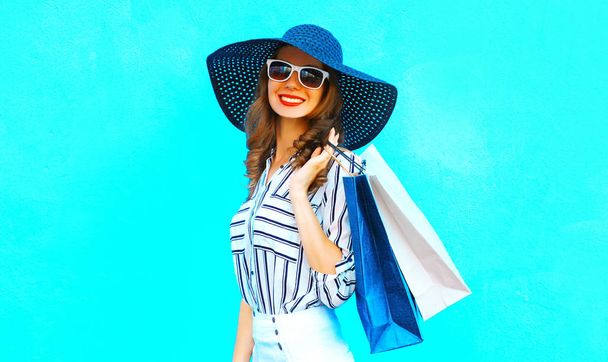Elegant young smiling woman wearing a shopping bags, straw hat, white pants over colorful blue background posing in the city  - Photo, Image