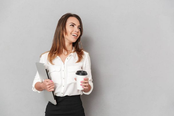 Portrait of a happy young business woman looking away while holding laptop computer and cup of coffee to go isolated over white background - Valokuva, kuva