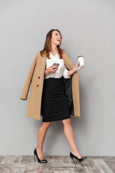 Image of happy young business woman standing isolated over grey wall background holding coffee chatting by phone. - Fotó, kép
