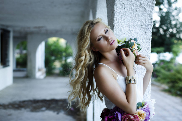 Młoda blond kobieta o niebieskich oczach, stojąc na ganku. Atrakcyjna dziewczyna z zamiarem i spokojny wygląd sobie romantyczną białą sukienkę i kwiaty - Zdjęcie, obraz