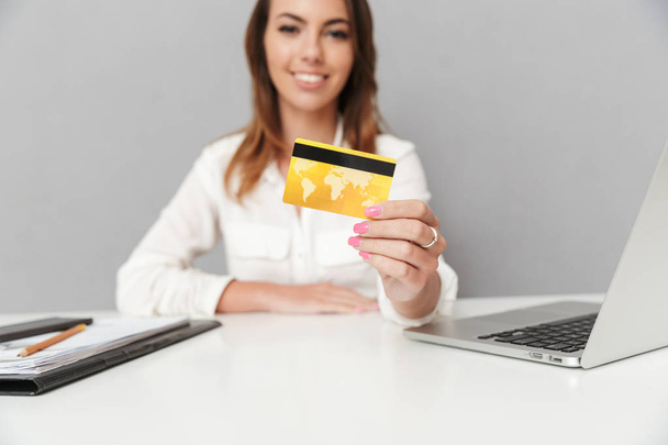 Close up of a happy young business woman showing golden credit card while sitting at the office table isolated over white background - Foto, Imagem