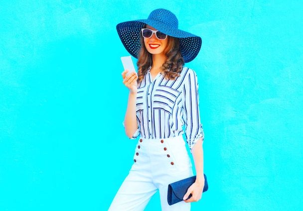 Happy girl is using a smartphone wearing a straw hat, white pants with a handbag clutch over colorful blue background  - 写真・画像