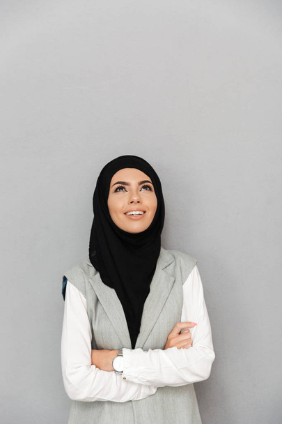 Απομονωμένη εικόνα της μουσουλμανικής κομψή γυναίκα 20s στο χιτζάμπ χαμογελώντας και κοιτάζοντας προς τα πάνω σε copyspace πάνω από το γκρίζο φόντο - Φωτογραφία, εικόνα