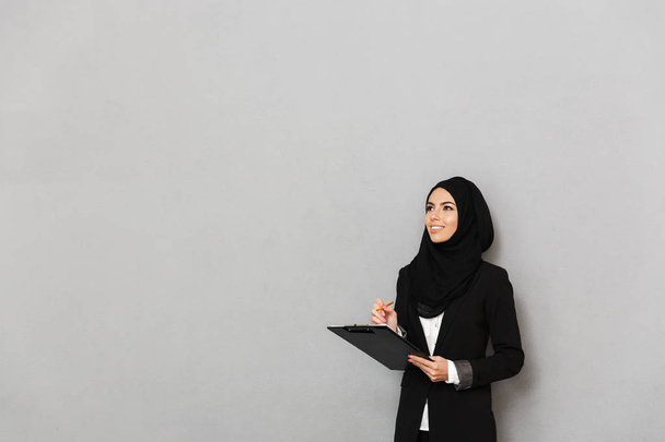 Portrait of a smiling young arabian woman holding clipboard with documents isolated over gray background - Foto, Imagem