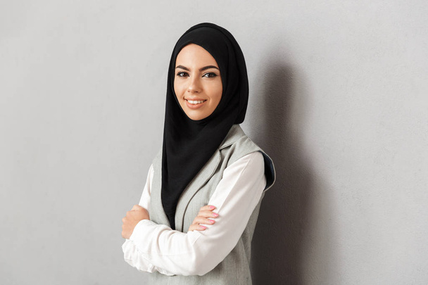 Muotokuva hymyilevä nuori arabialainen nainen seisoo kädet taitettuna ja katsot kameraa eristetty yli harmaa tausta
 - Valokuva, kuva