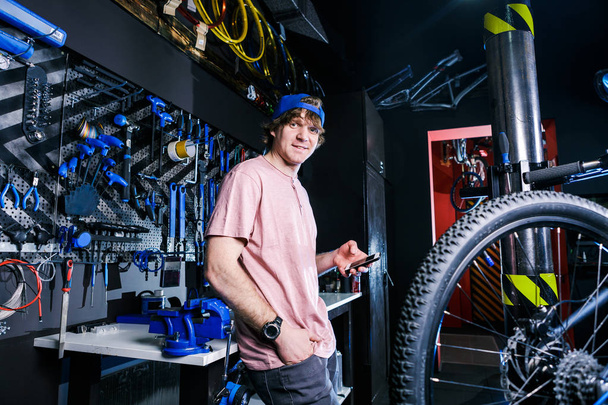 Een jonge, modieuze roodharige mannelijke kleine ondernemer verkopen en repareren van een fiets is het dragen van een blauwe cap en een roze trui is het gebruik van een mobiele telefoon terwijl je in een fietswinkel. - Foto, afbeelding