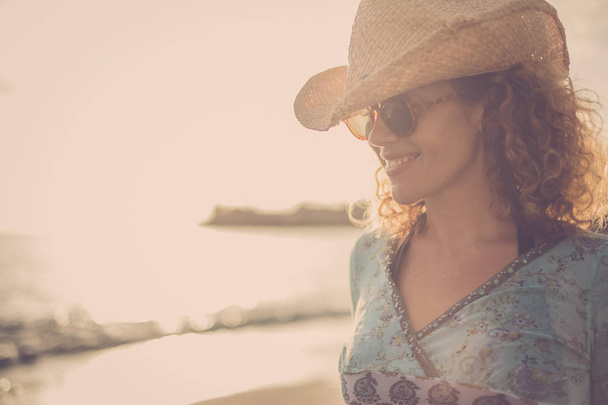 krásné a úžasné blond kudrnaté vlasy modelu středního věku s úsměvem kovbojský klobouk a pozice na pláži s zlatý západ slunce v pozadí. barvy a svobody nezávislosti silná žena koncept - Fotografie, Obrázek
