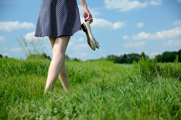 Female walks on the grass with barefoot. Young woman enjoys outdoor walk, nature, green fields and blue sky. - Фото, изображение