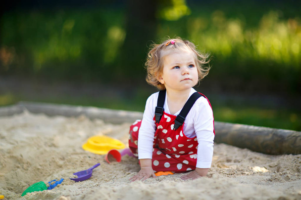Χαριτωμένο μικρό παιδί κορίτσι παίζει στην άμμο σε υπαίθρια παιδική χαρά. Όμορφο μωρό σε κόκκινο χρώμα ούλων παντελόνι έχοντας διασκέδαση στην ηλιόλουστη ζεστή καλοκαιρινή μέρα. - Φωτογραφία, εικόνα