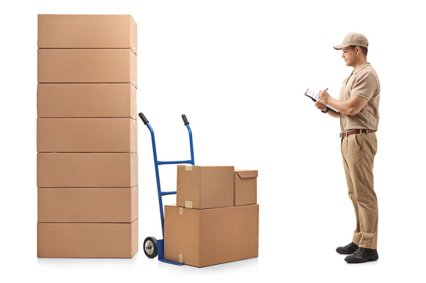 Full length profile shot of a delivery man standing next to a hand truck and a stack of boxes writing in a clipboard isolated on white background - Fotografie, Obrázek