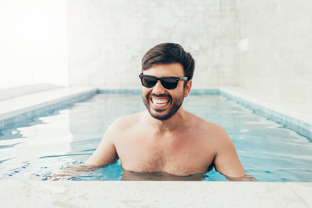 Close up portrait of attractive young man in sunglasses resting on edge of swimming pool - Foto, imagen