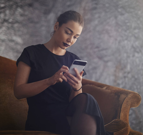Young attractive girl holding mobile phone and texting message while sitting on the couch - Foto, Imagem