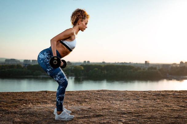 Young fitness woman doing workout with dumbbells by river at sunset - Photo, Image