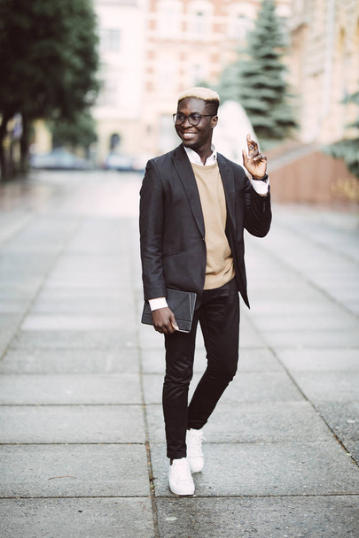 Handsome young Afro American businessman in sunglasses is holding on the tablet walking in the street - Fotoğraf, Görsel