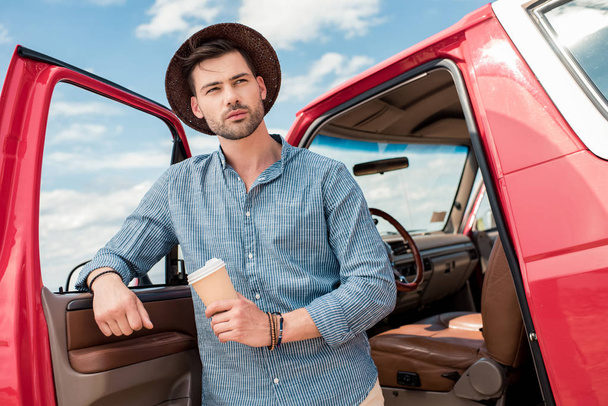 handsome man in hat holding coffee to go and standing at car during road trip - Foto, Imagem
