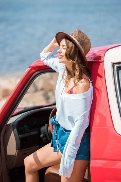 attractive woman in hat posing at red car during road trip near the sea - Foto, Imagen