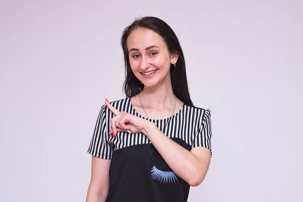 Look Here There is something interesting. Portrait of a beautiful brunette girl in a dress on a white background showing on the product. She is standing right in front of the camera smiling and looking happy - Fotó, kép