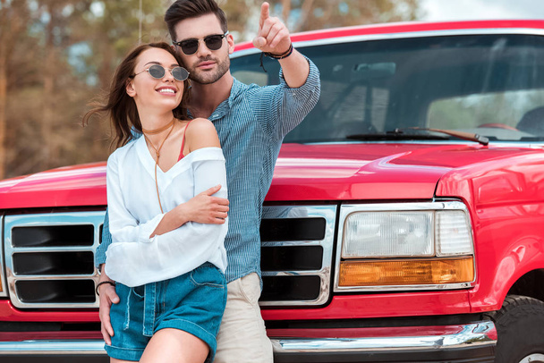 smiling man hugging his girlfriend and pointing somewhere while standing near red jeep - Foto, imagen
