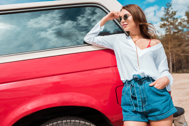 aantrekkelijk meisje in zonnebril poseren in de buurt van rode auto tijdens de reis - Foto, afbeelding