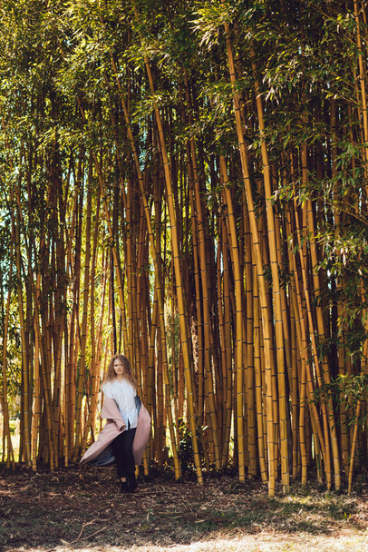 krullend elegante jonge meisje in een lang roze jas wandelingen in een park naast Bamboe bomen, geniet van de ontspannen - Foto, afbeelding