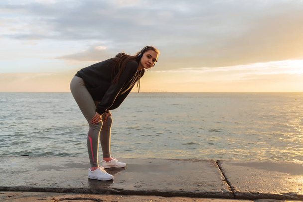 a cute young girl in sportswear jumped out by the sea at sunset, listening to music on headphones - Foto, Imagem