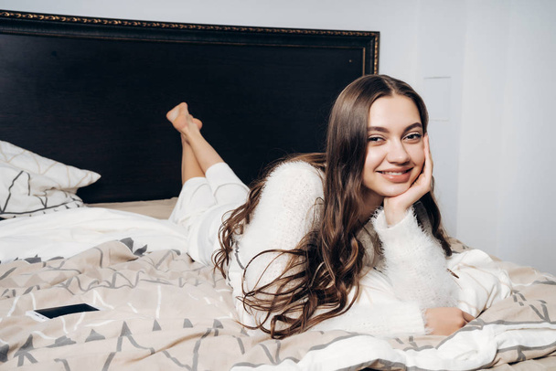 happy long-haired girl in pajamas lies on a bed early in the morning, in a good mood, smiling - Fotó, kép